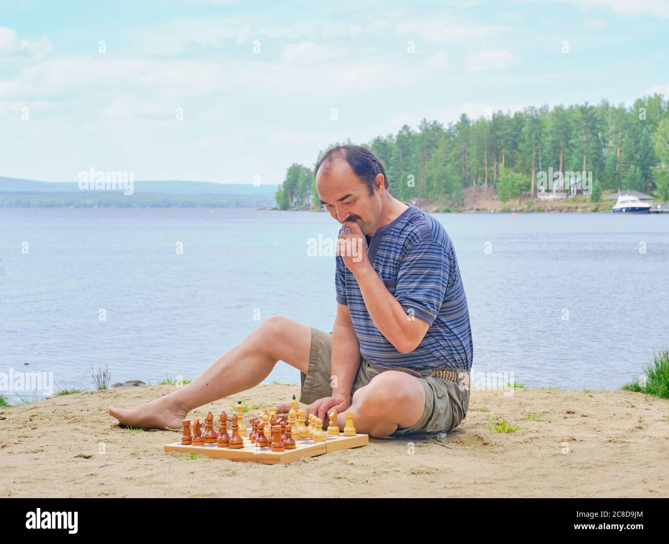 Homme asiatique senior qui réfléchit à un mouvement dans une partie d'échecs et qui s'assoit sur la plage près du lac. Concept de détox numérique Banque D'Images