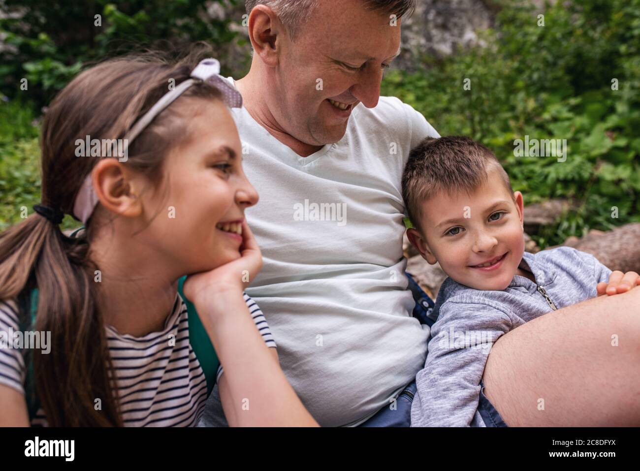 Un père souriant et ses deux enfants adorables assis sur un sentier faisant une pause dans leur randonnée en famille Banque D'Images