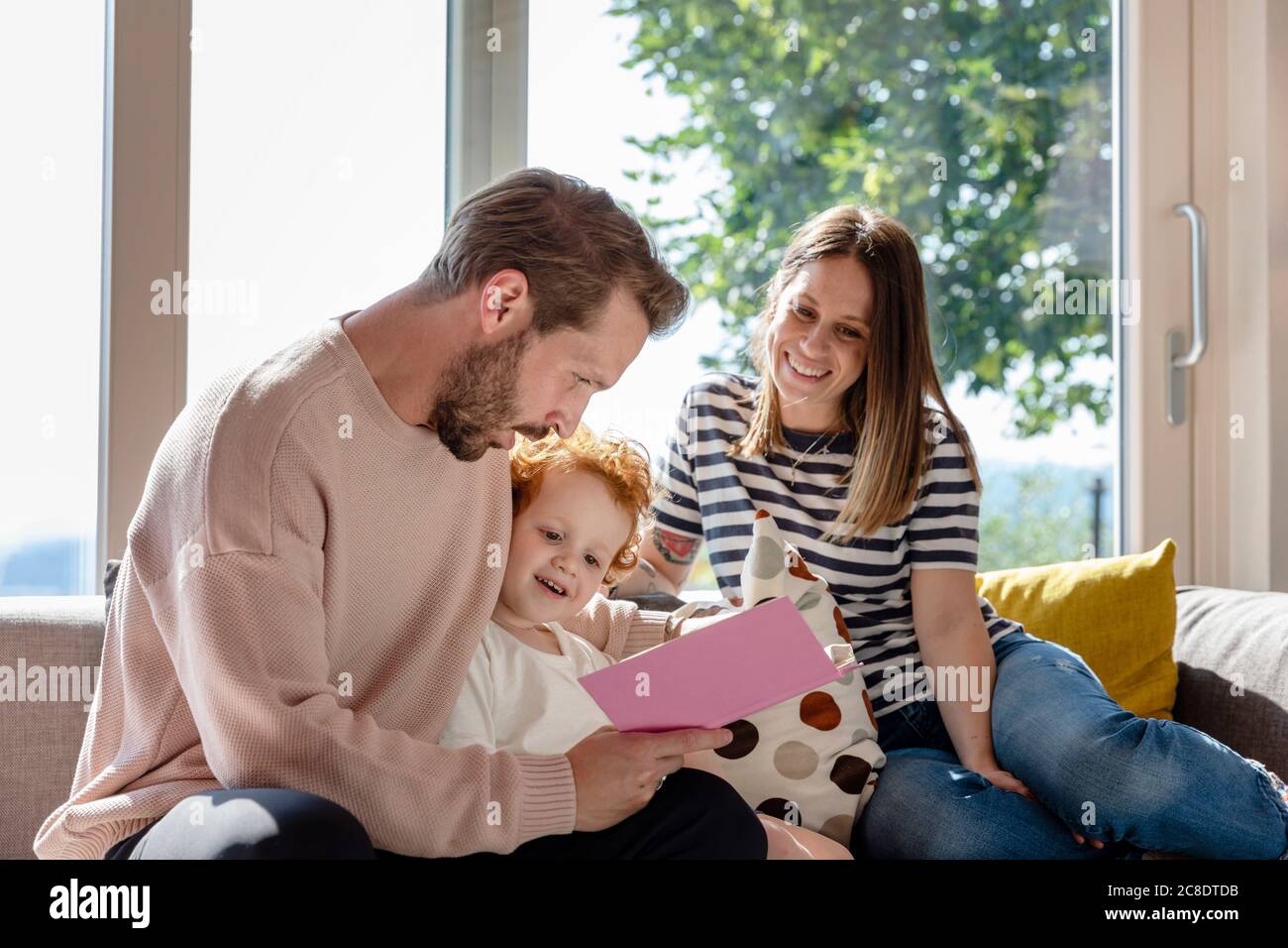 Femme souriante assise et regardant un homme lisant un livre d'images pour garçon dans la salle de séjour Banque D'Images