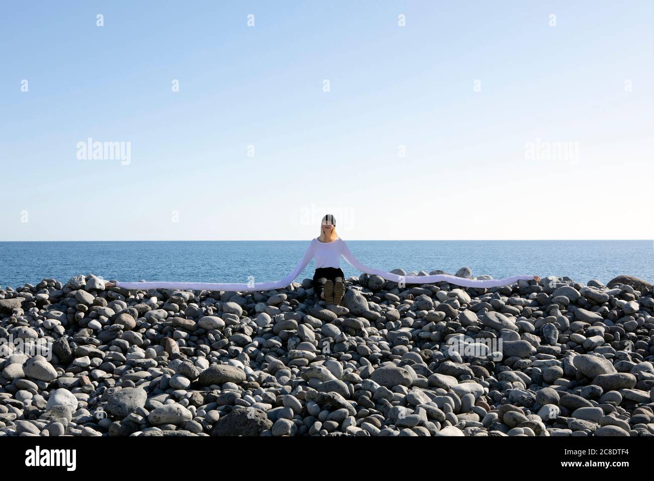 Femme aux longues mains artificielles assise sur des galets à la plage contre le ciel dégagé Banque D'Images