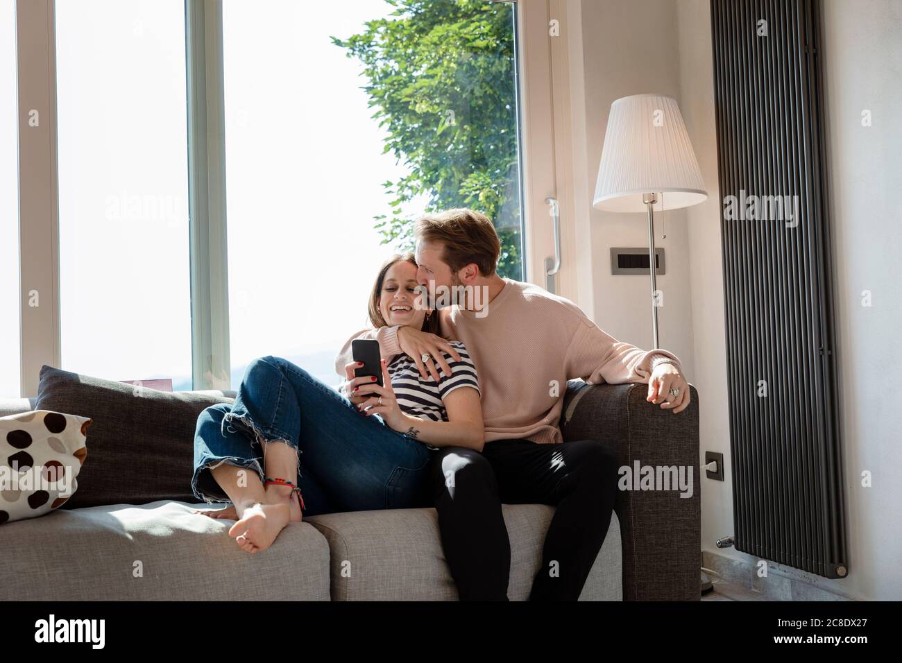 Homme embrassant la femme tout en se relaxant sur le canapé contre la fenêtre dedans salle de séjour à la maison Banque D'Images