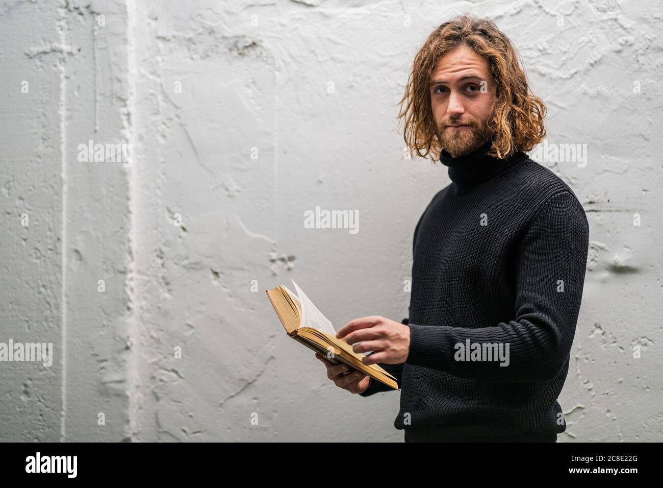 Jeune homme barbu tenant le livre en se tenant contre l'ancien mur à la maison Banque D'Images