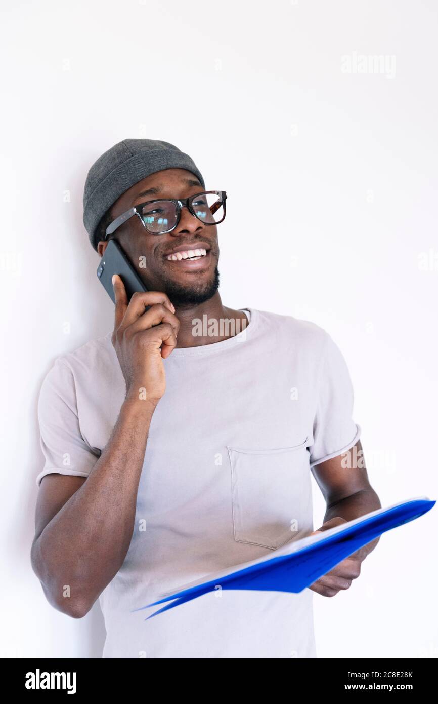 Jeune homme d'affaires souriant tenant des dossiers et parlant sur un smartphone tout en regardant loin sur fond blanc Banque D'Images