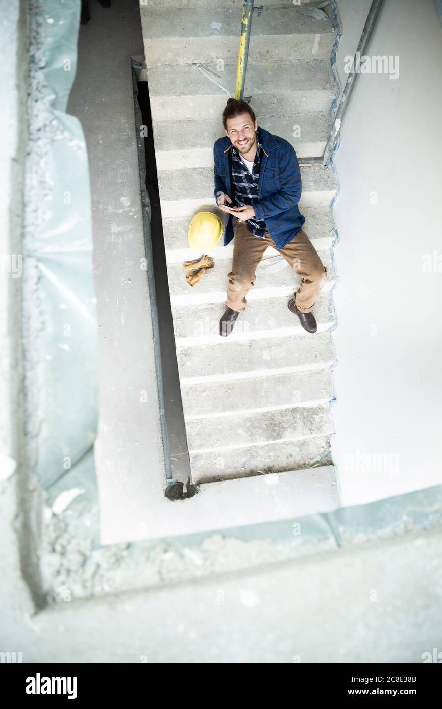 Jeune ouvrier souriant en construction utilisant un smartphone tout en étant assis sur des marches dans la construction de la maison Banque D'Images