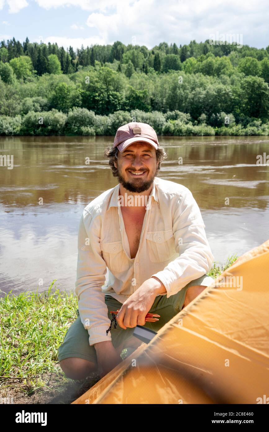 Homme souriant portant un chapeau installant une tente contre le lac au camping en forêt Banque D'Images