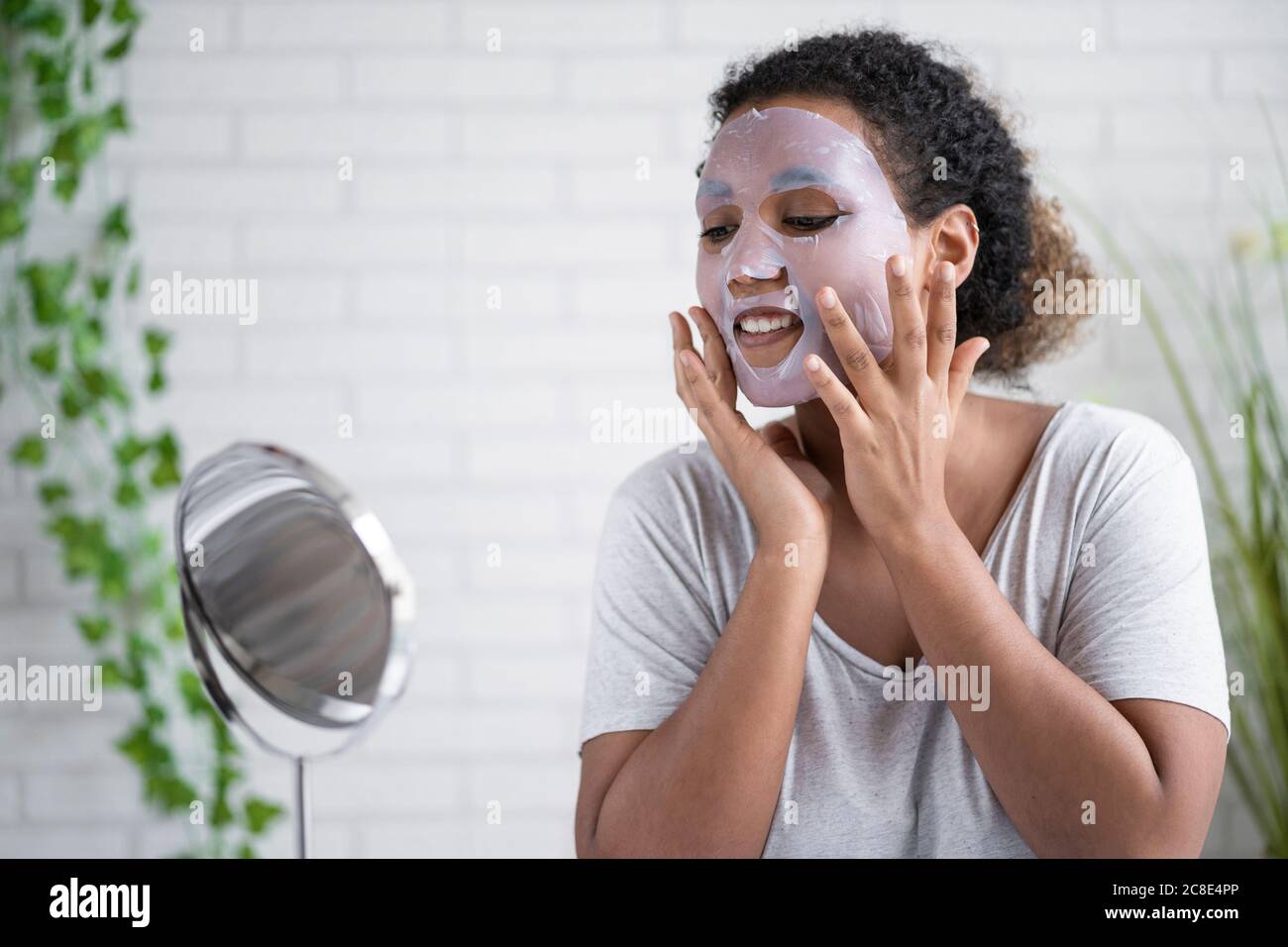 Jeune femme appliquant le masque facial en papier tout en regardant dans le miroir à la maison Banque D'Images