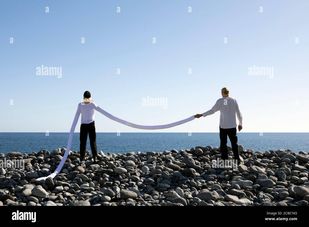 Femme aux bras longs artificiels tenant la main de l'homme sur des cailloux à la plage contre un ciel dégagé Banque D'Images