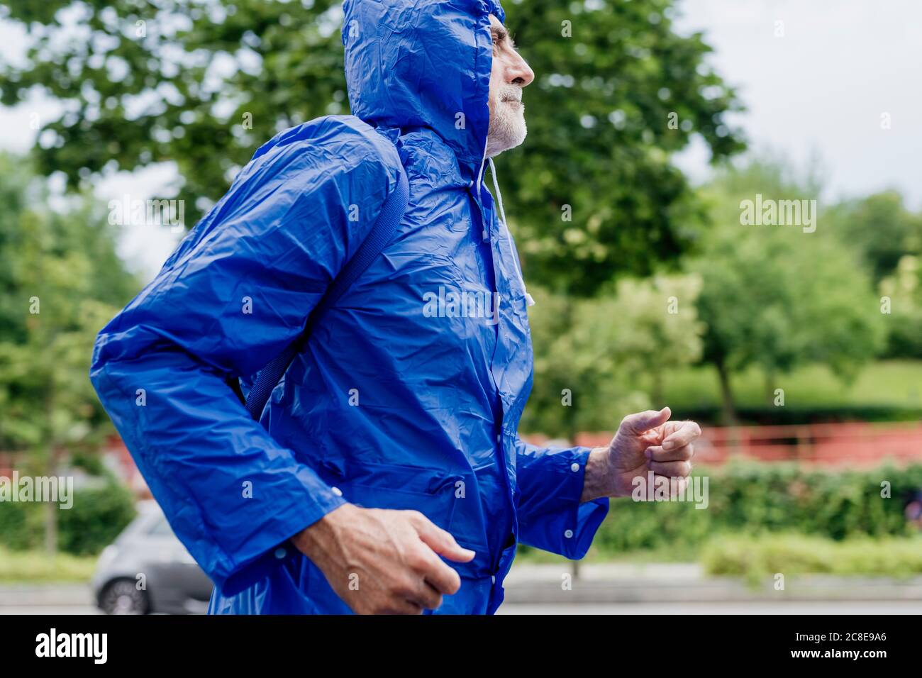 Homme âgé portant un imperméable bleu qui court contre les arbres dans le parc Banque D'Images