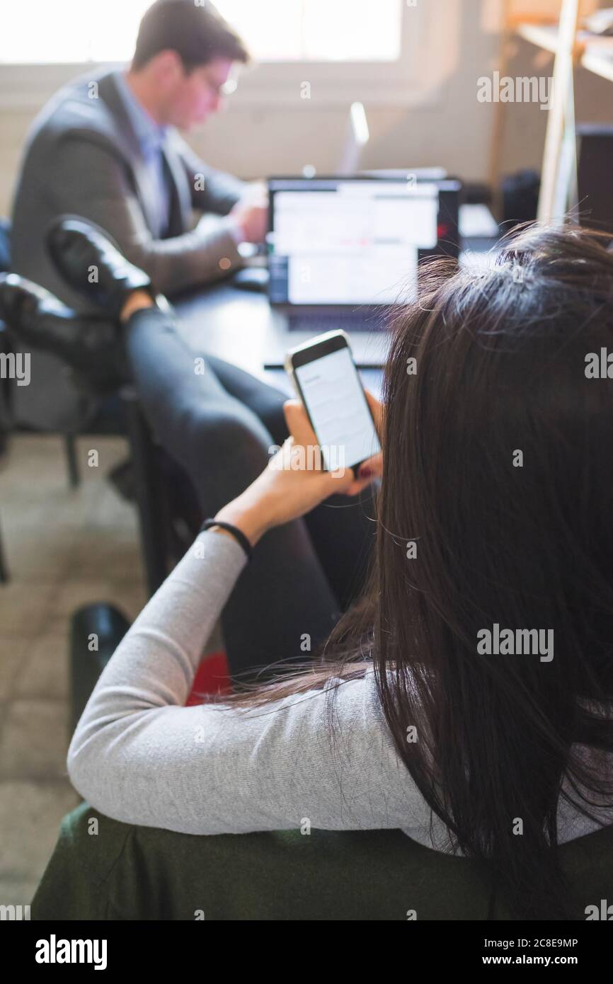 Femme détendue utilisant un smartphone au bureau avec un homme d'affaires en arrière-plan en fonctionnement Banque D'Images