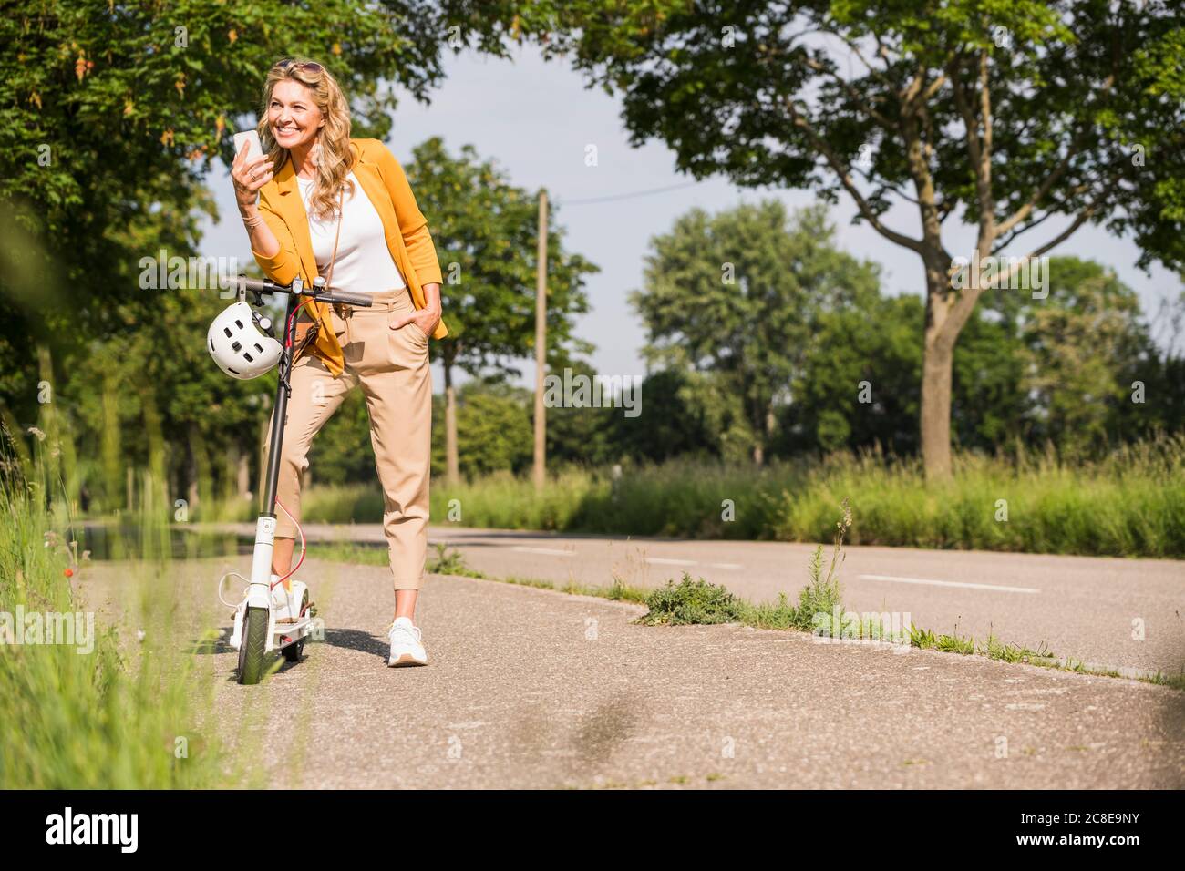 Jeune femme à la mode tenant un smartphone debout avec une pression scooter sur trottoir Banque D'Images