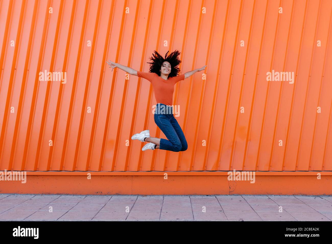 Bonne jeune femme sautant avec les bras tendus contre le mur orange Banque D'Images
