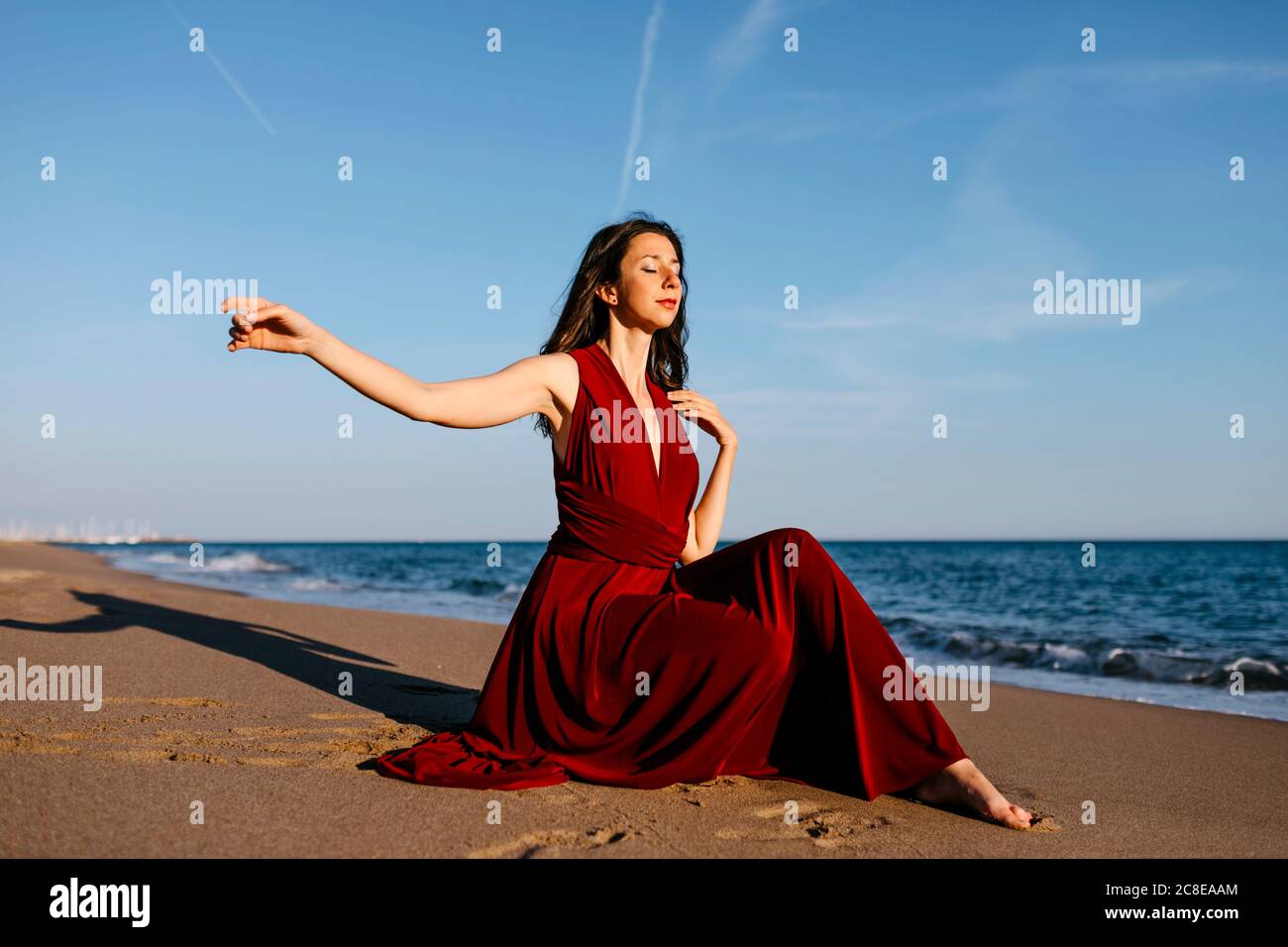 Femme délicate en robe rouge sittingh sur la plage, sentant le soleil Banque D'Images