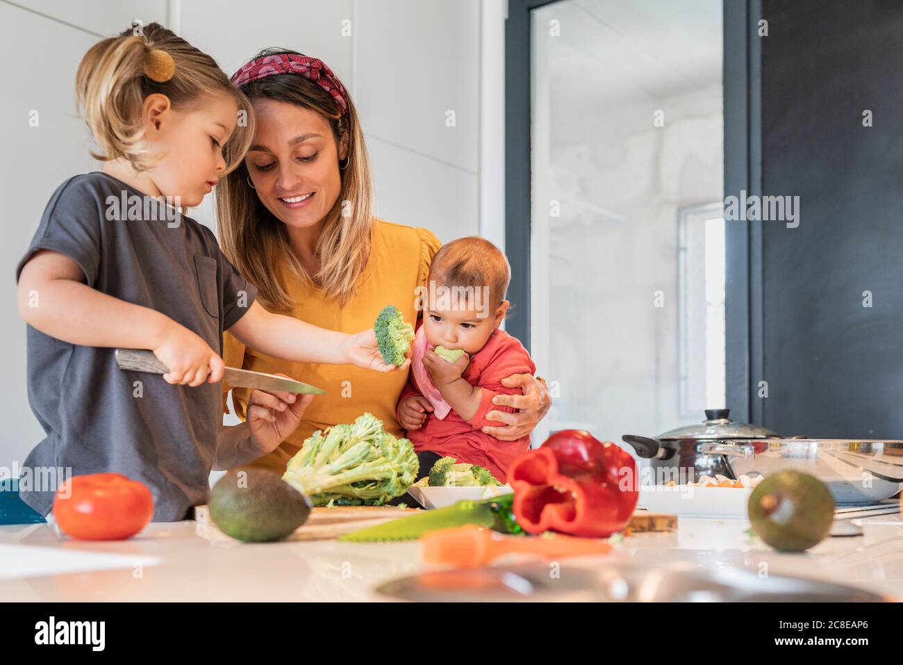 Mère tenant la petite fille tout en guidant la fille dans la coupe de légumes sur l'île de cuisine Banque D'Images