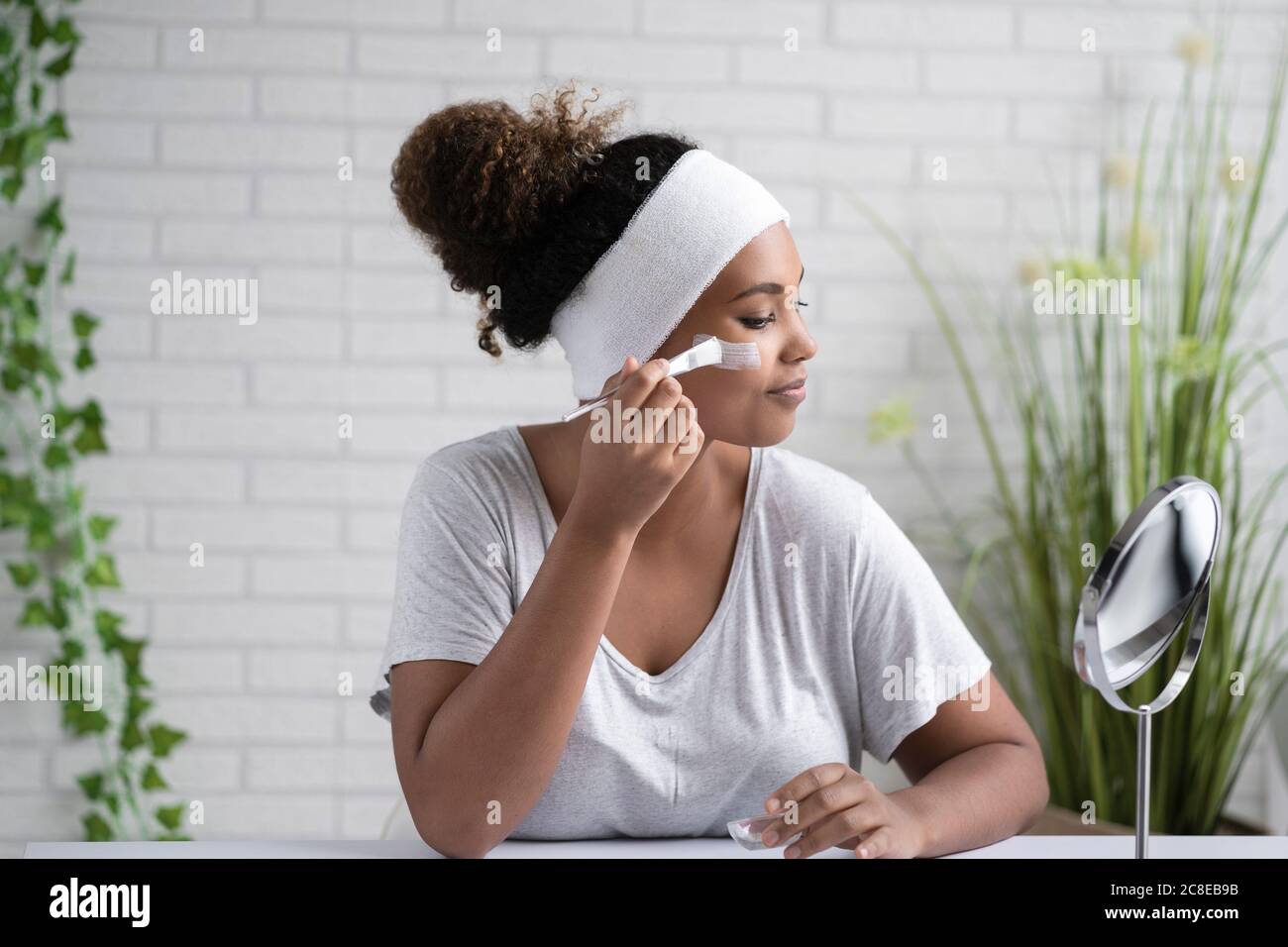 Jeune femme portant un serre-tête appliquant un masque facial tout en regardant à l'intérieur miroir à la maison Banque D'Images