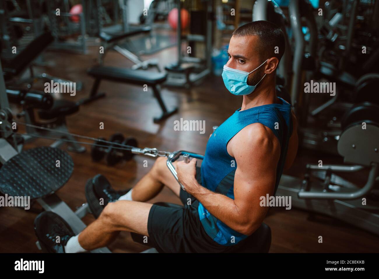Jeune homme de forme musculaire avec masque médical faisant un exercice lourd de poids pour le dos et le bras sur la machine dans la salle de gym.prévention de COVID - 19 coronavirus Banque D'Images