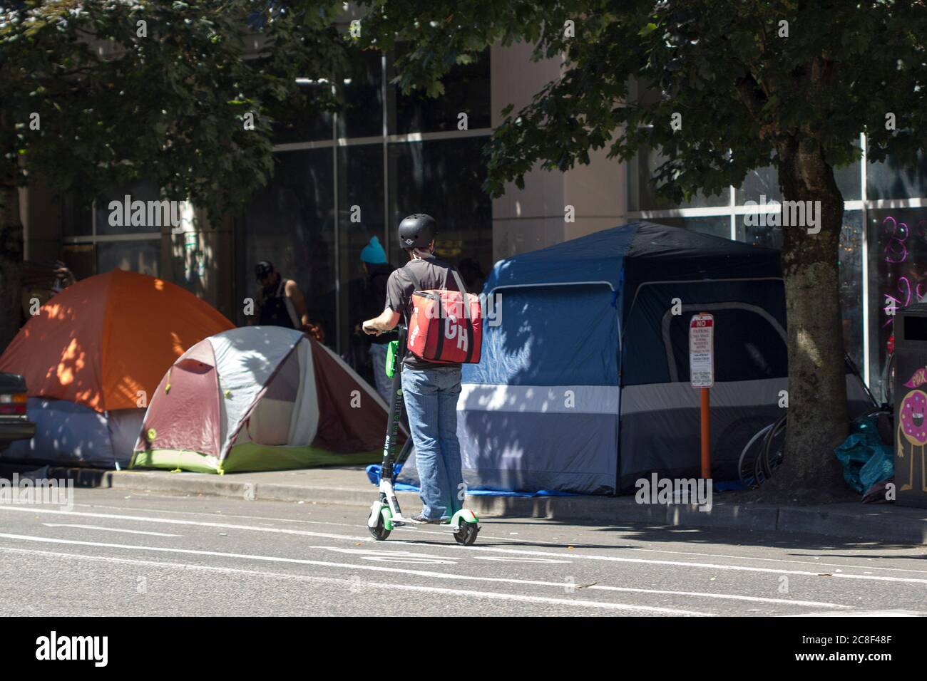 Un employé de Grubhub qui a un sac fourre-tout se déplace sur un scooter électrique le long d'une rue bordée de tentes sans abri dans le centre-ville de Portland pendant la pandémie de COVID. Banque D'Images