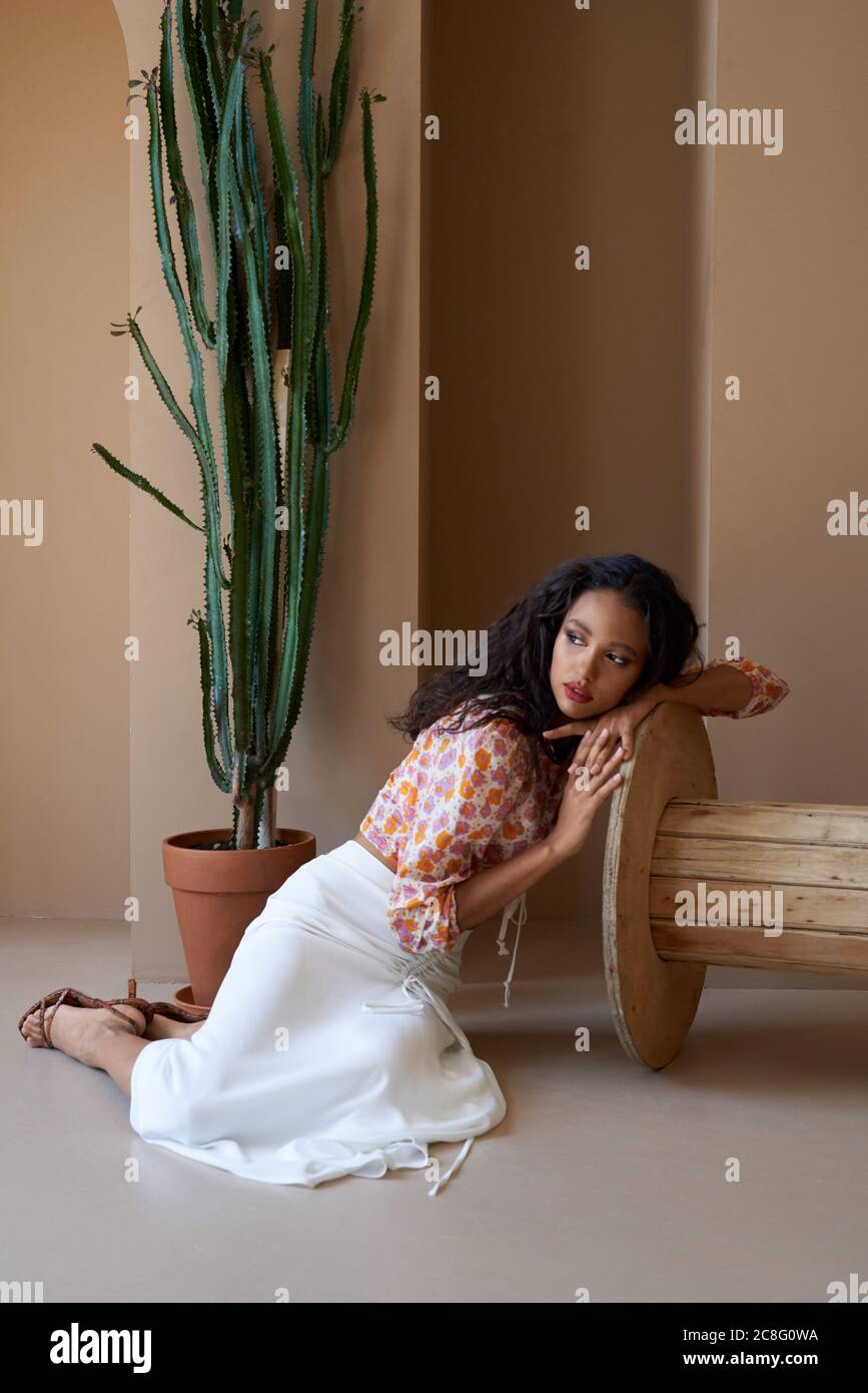 Magnifique mulatto fille avec des cheveux bouclés posant près de grande plante succulente dans le pot de fleur, assis sur le sol et s'appuyant sur la roue en bois. Superbe femme à moitié africaine portant un chemisier, une jupe blanche et des talons. Banque D'Images