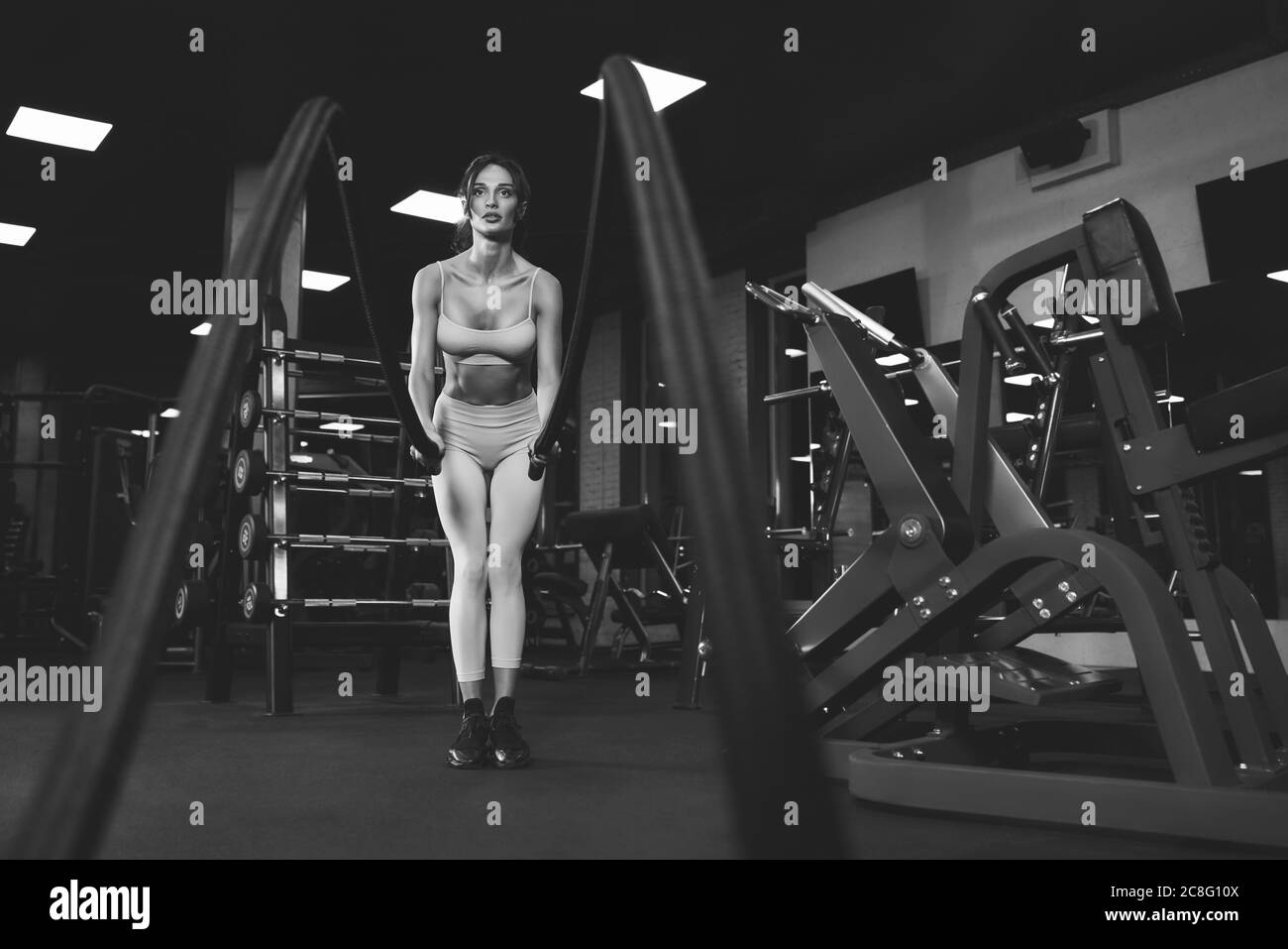 De dessous de vue de la femme brune en sport léger entraînement dans la salle de gym vide avec cordes. Portrait monochrome pleine longueur de la belle fitnesswoman avec le visage fort faisant cardio. Concept de sport. Banque D'Images