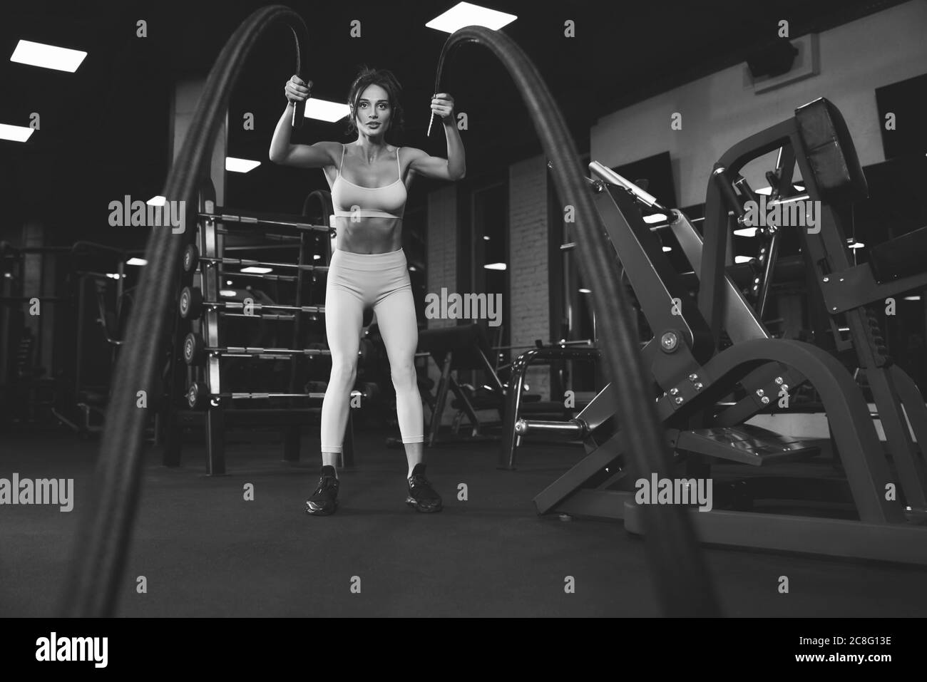 De dessous de vue de la femme brune en sport léger entraînement dans la salle de gym vide avec cordes. Portrait monochrome pleine longueur de la belle fitnesswoman avec le visage fort faisant cardio. Concept de sport. Banque D'Images