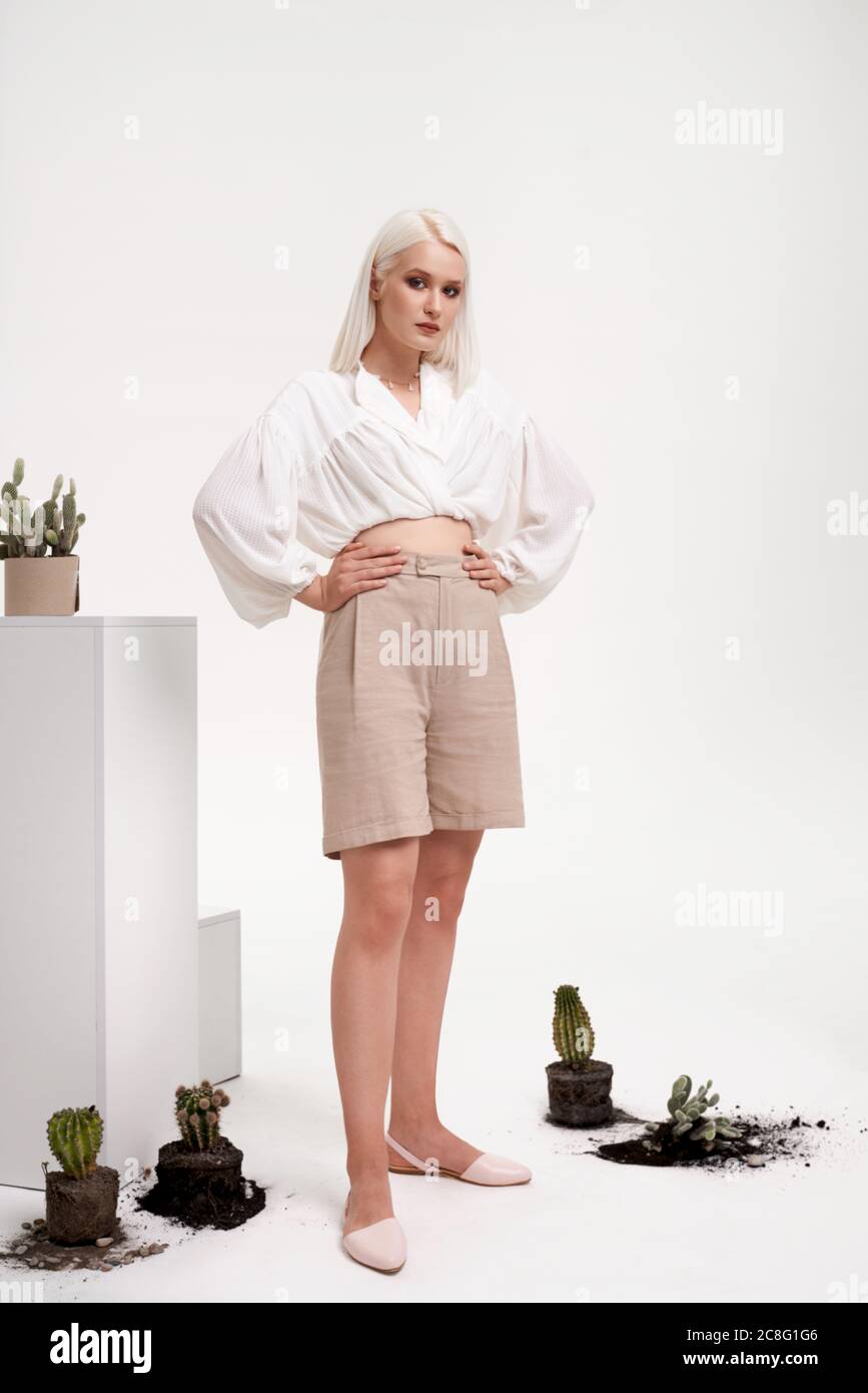 Portrait d'une fille portant une tenue d'été élégante posant près des boîtes et des cactus avec le sol sans pots isolés sur blanc. Jeune modèle blond tenant les mains à la taille et regardant la caméra. Banque D'Images