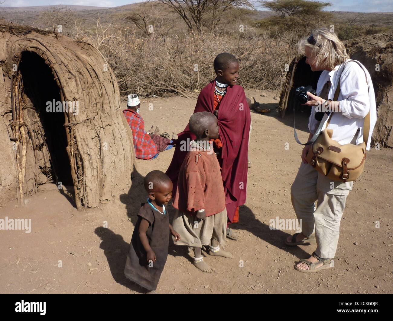Sympathique, blonde, femme touriste avec caméra à la main discutant avec trois enfants timides et souriants Masai dans un village tribal avec des huttes de boue, Tanzanie. Banque D'Images