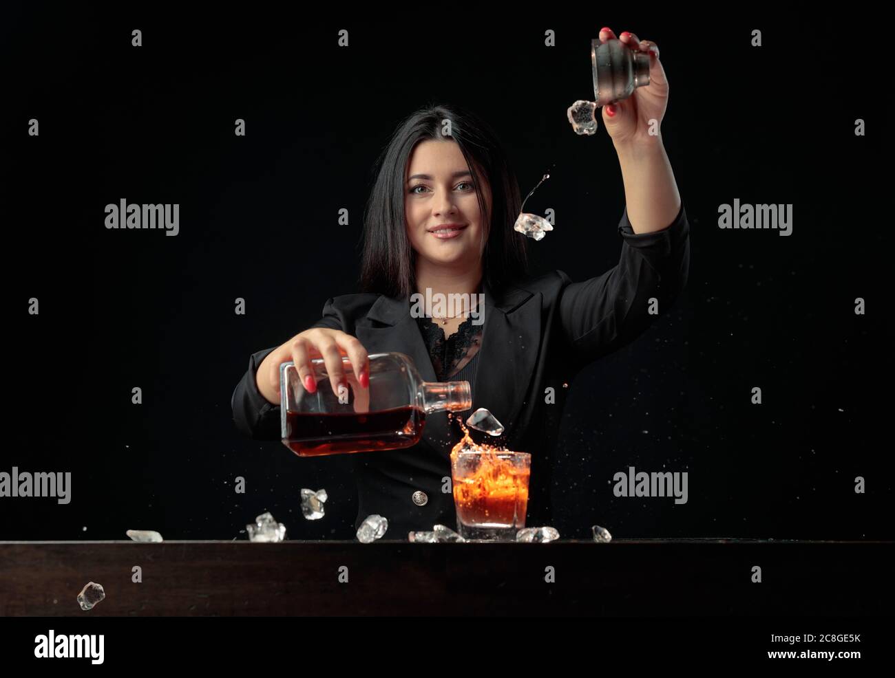 Une belle barman verse une boisson alcoolisée dans un verre et jette des morceaux de glace. Magnifique brunette en noir préparer un cocktail. Banque D'Images