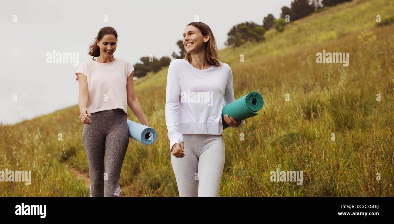 Des femmes qui vont à l'extérieur pour s'entraîner. Deux femmes gaies marchant dans les champs portant des tapis de yoga. Banque D'Images