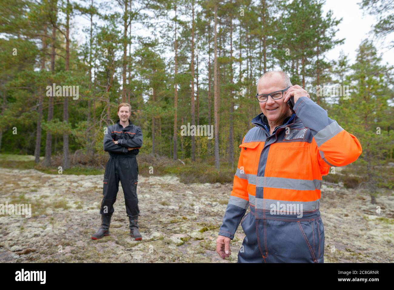 Heureux homme mature parlant sur téléphone mobile dans la forêt avec heureux jeune homme dans l'arrière-plan Banque D'Images