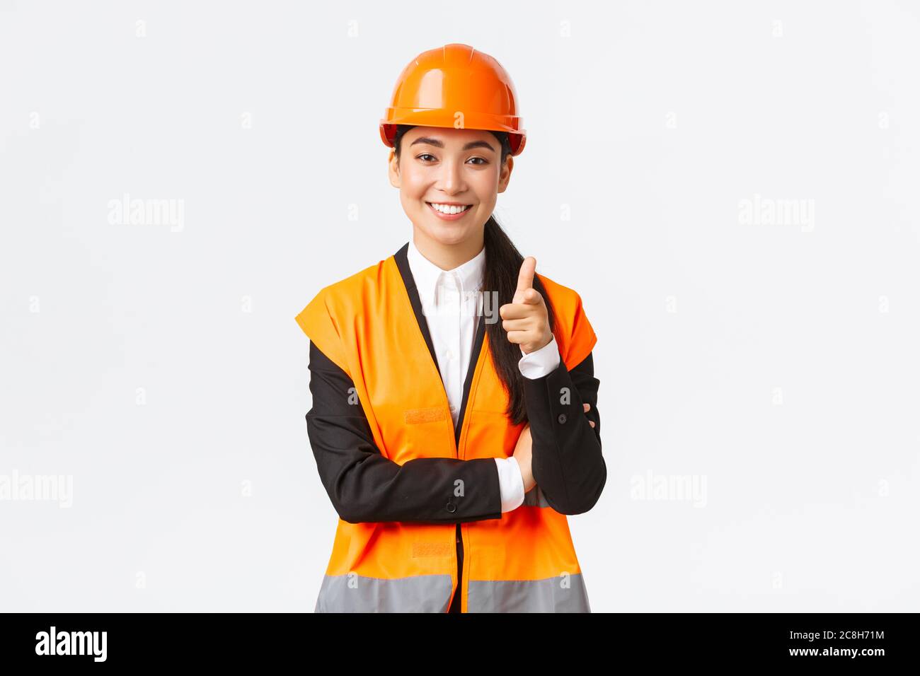 Une femme asiatique souriante et réussie, qui pointe le doigt sur la caméra, porte un casque de sécurité et une veste réfléchissante, présente la construction Banque D'Images