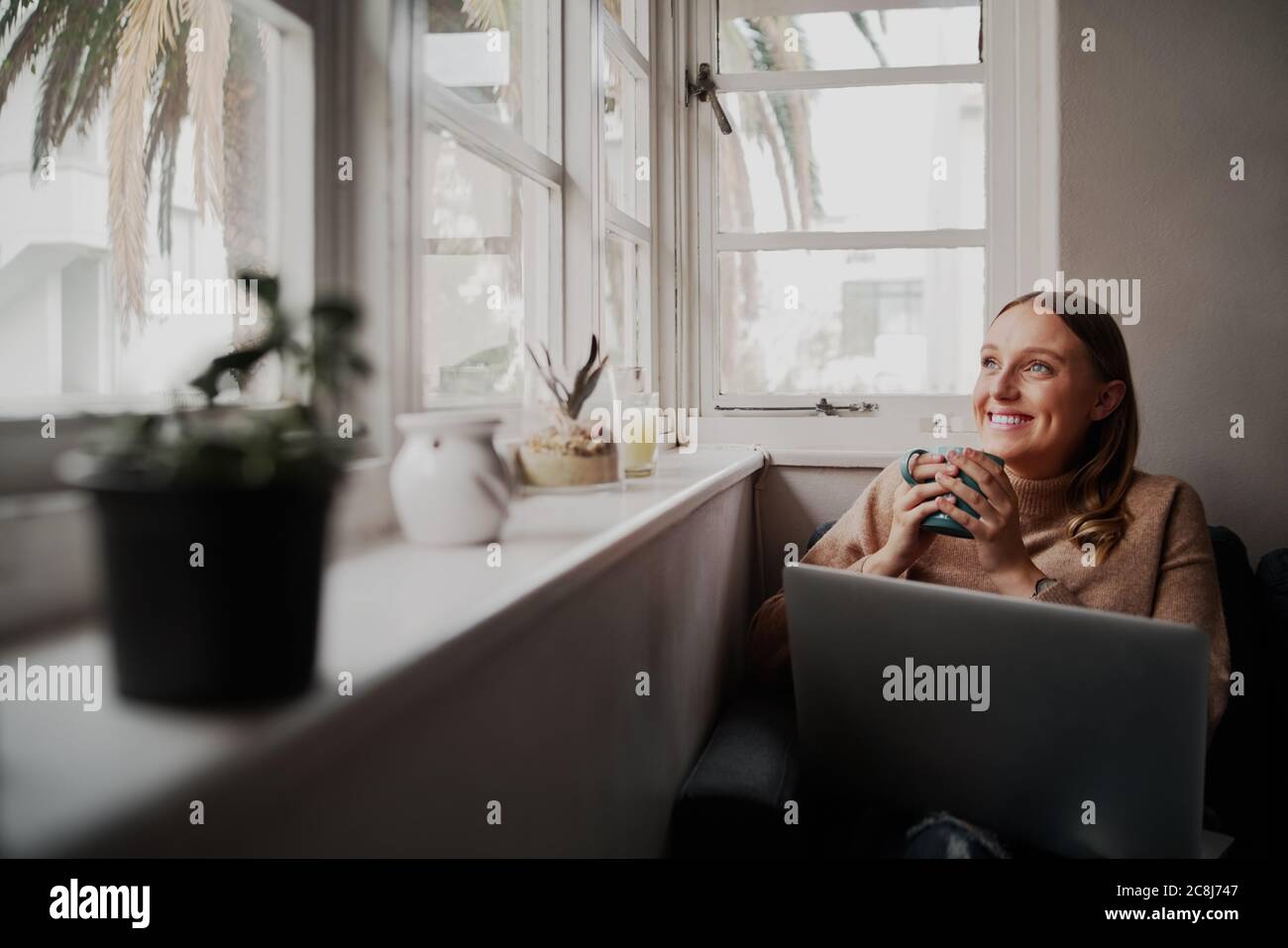 Jeune femme d'affaires réussie utilisant un ordinateur portable à la maison pendant la quarantaine tenant une tasse de café dans la main tout en regardant par la fenêtre Banque D'Images