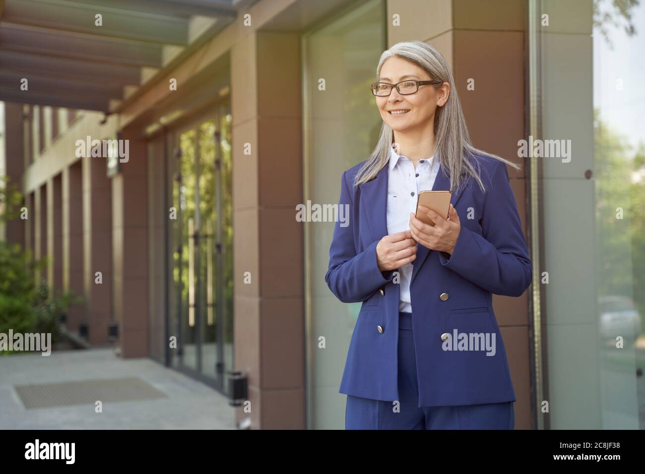 Bonne femme d'affaires mûre et attrayante en costume classique utilisant un smartphone et souriant tout en se tenant contre le bâtiment de bureau à l'extérieur. Professionnels Banque D'Images