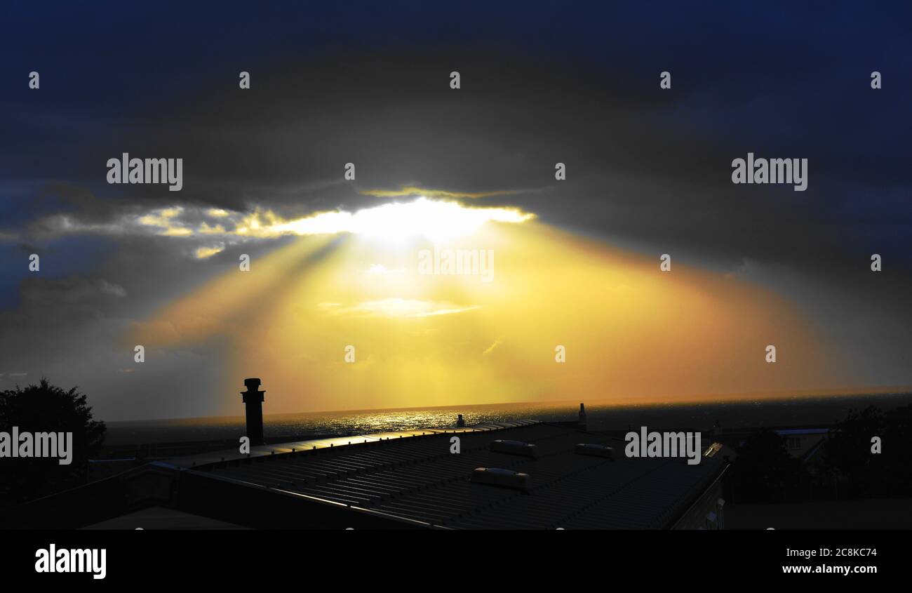 Des puits de lumière du soleil se brisent à travers un trou dans les nuages sombres tandis que le soleil se couche sur l'horizon. Banque D'Images
