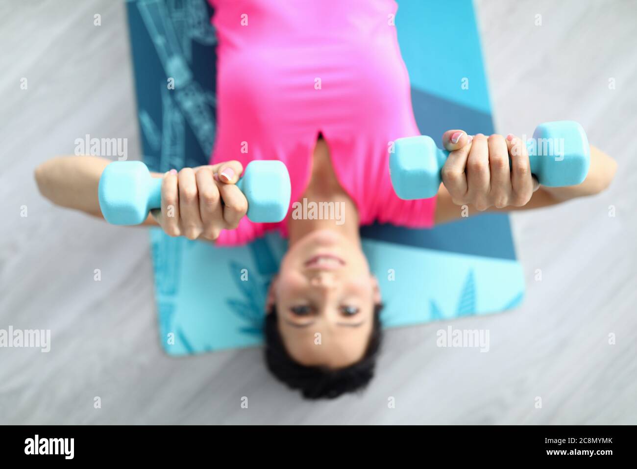 Belle femme en T-shirt rose s'allonger sur le tapis de gymnastique et rais ses bras avec des haltères au-dessus d'elle-même. Banque D'Images