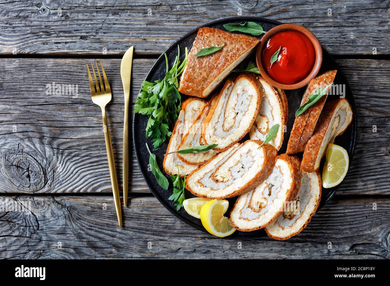Omelette au fromage rouler avec garniture de viande hachée sur une assiette noire avec citron, ketchup et persil sur une table en bois gris avec fourchette dorée et couteau, vertic Banque D'Images