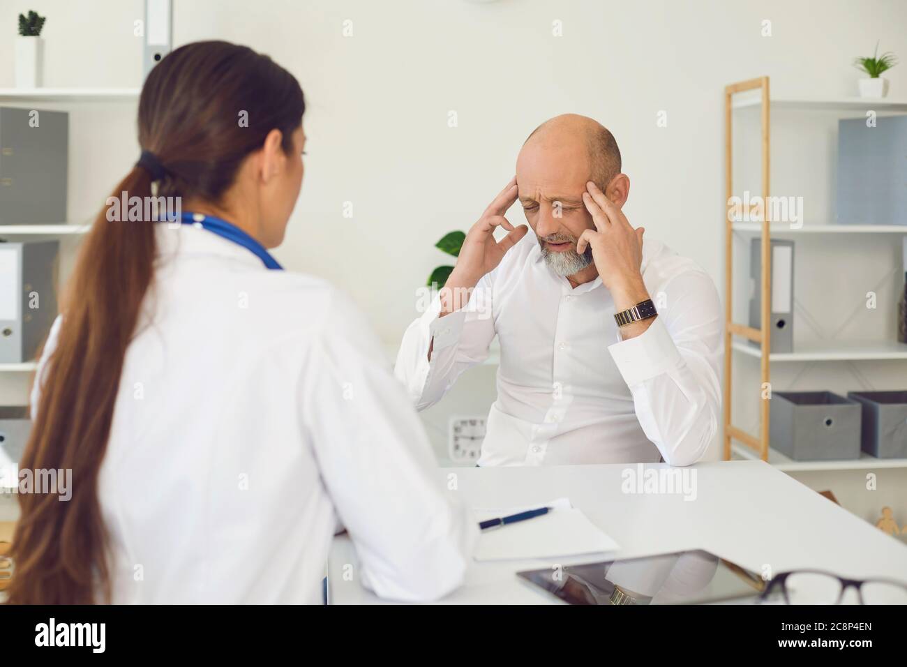 Maux de tête. Le senor du patient indique au médecin les symptômes de maux de tête assis sur une chaise dans un bureau de clinique. Banque D'Images
