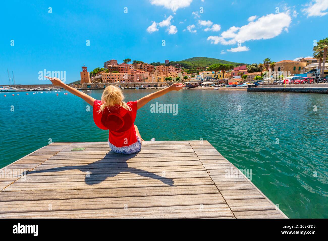 Femme touristique assise sur la jetée et regardant le port de Rio Marina de l'île d'Elbe en costume rouge. Style de vie touriste fille sur le voyage de vacances en Italie Banque D'Images