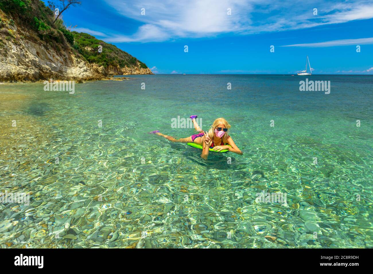 Femme avec le signe de victoire main sur matelas flottant de plage avec un masque chirurgical rose pendant Covid-19. Femme nageant à Prunini o Seccione Beach dans Banque D'Images