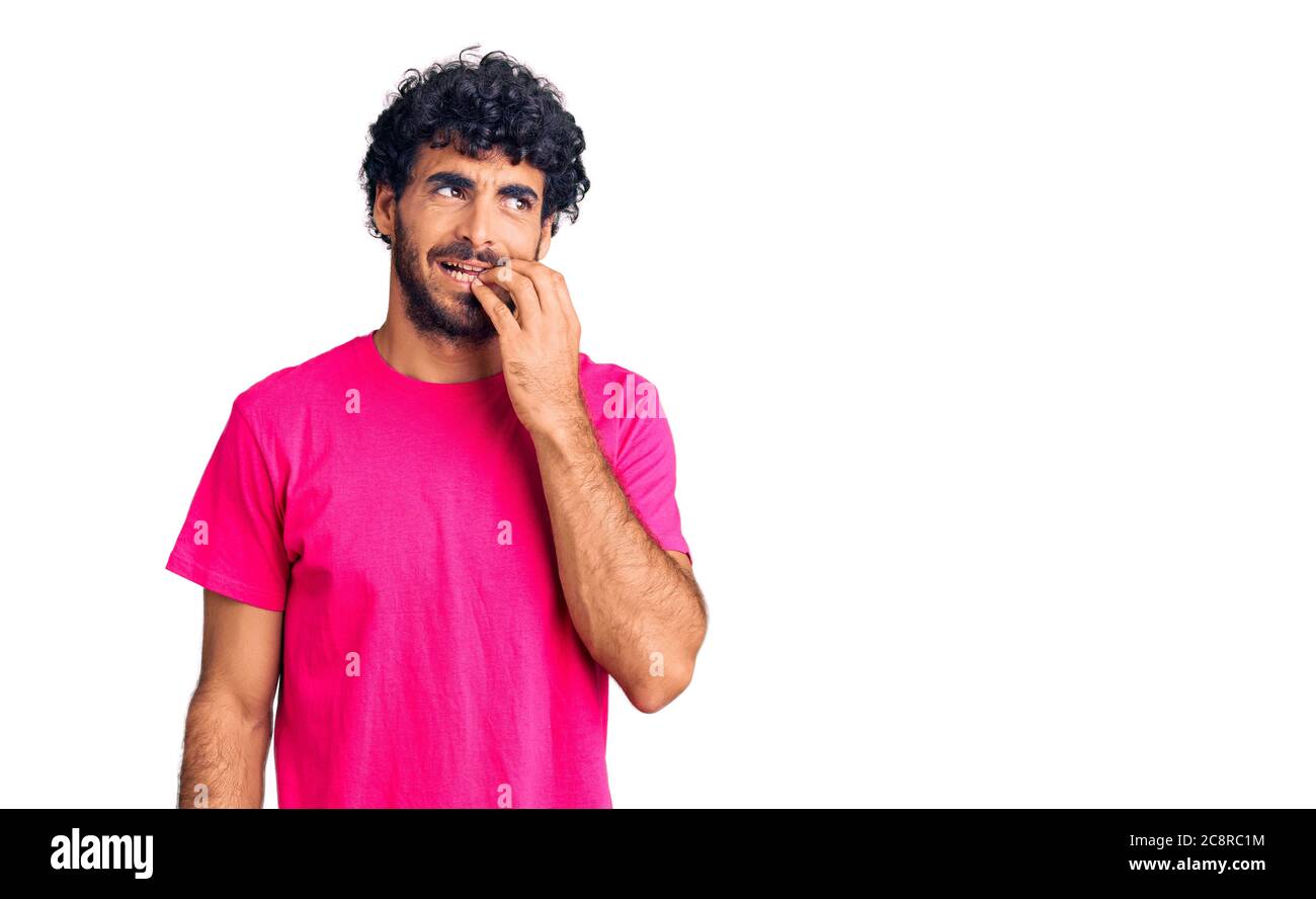 Beau jeune homme aux cheveux bouclés et ours portant un t-shirt rose décontracté, il a l'air stressé et nerveux avec les mains sur la bouche piquant les ongles. Problème d'anxiété. Banque D'Images