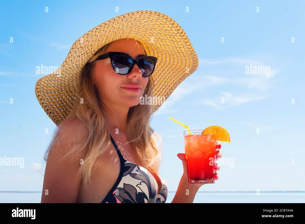 Une jeune femme en lunettes de soleil et un chapeau de paille tient un  cocktail dans sa main contre le ciel bleu et la mer. Bonheur fille porter  bikini à la plage