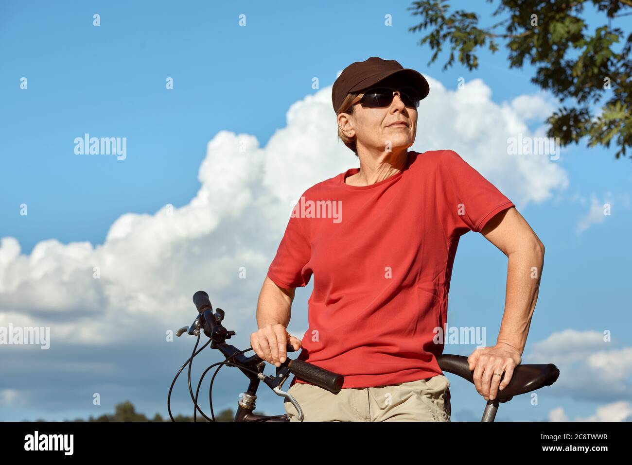 Une femme d'âge moyen dans un T-shirt rouge et une casquette repose près du vélo et profite du beau temps ensoleillé. Banque D'Images