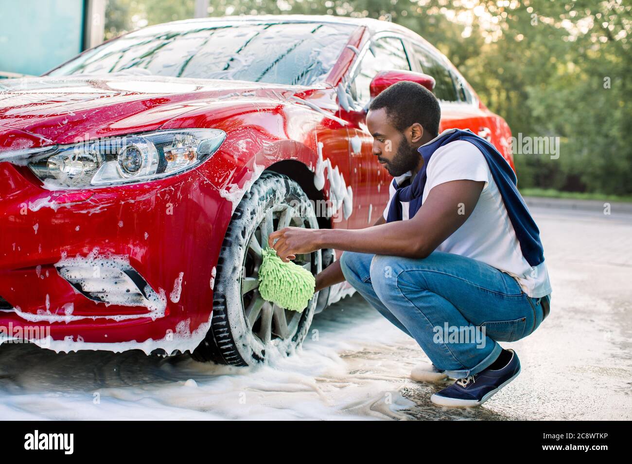 Voiture rouge de luxe dans la mousse de savon dehors au service de lavage de  voiture. Vue latérale du jeune homme africain charmant utilisant un gant de  lavage de voiture en microfibre