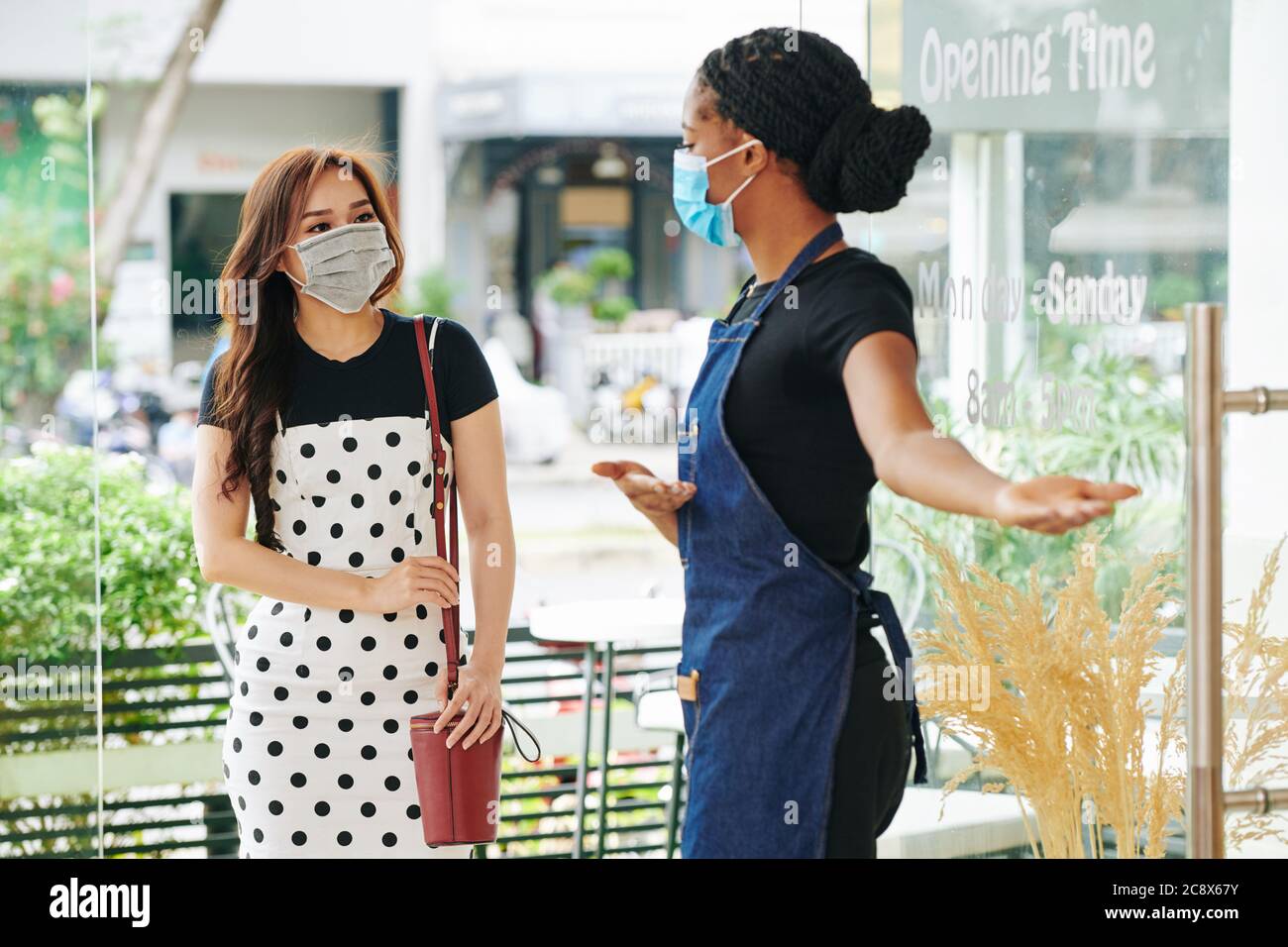 Serveuse en tablier et masque médical faisant un geste d'accueil quand invitant une jolie jeune femme Banque D'Images