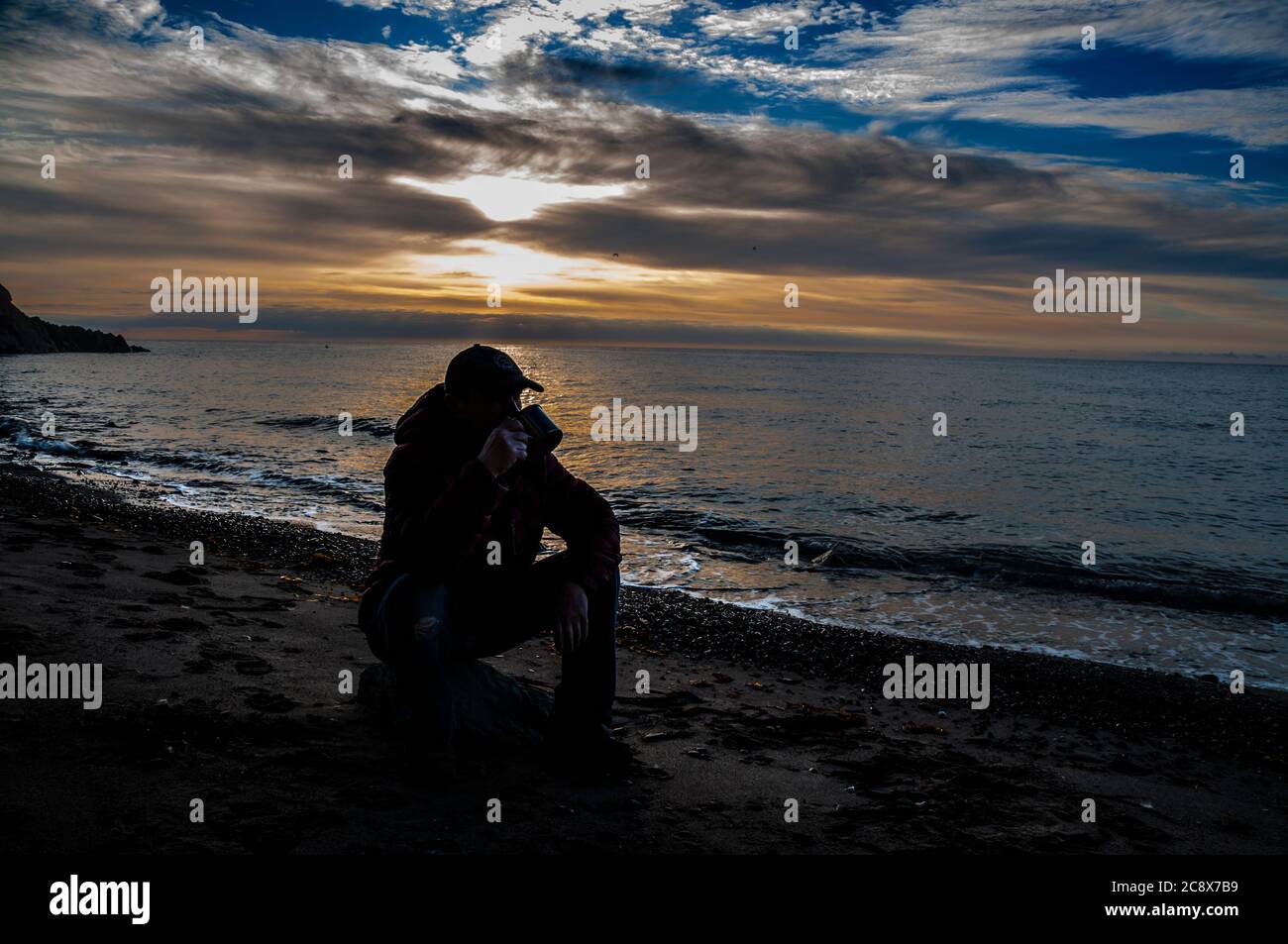 Tasse de thé le matin. Homme assis sur un rocher et buvant du thé au lever du soleil sur une plage de mer. Banque D'Images