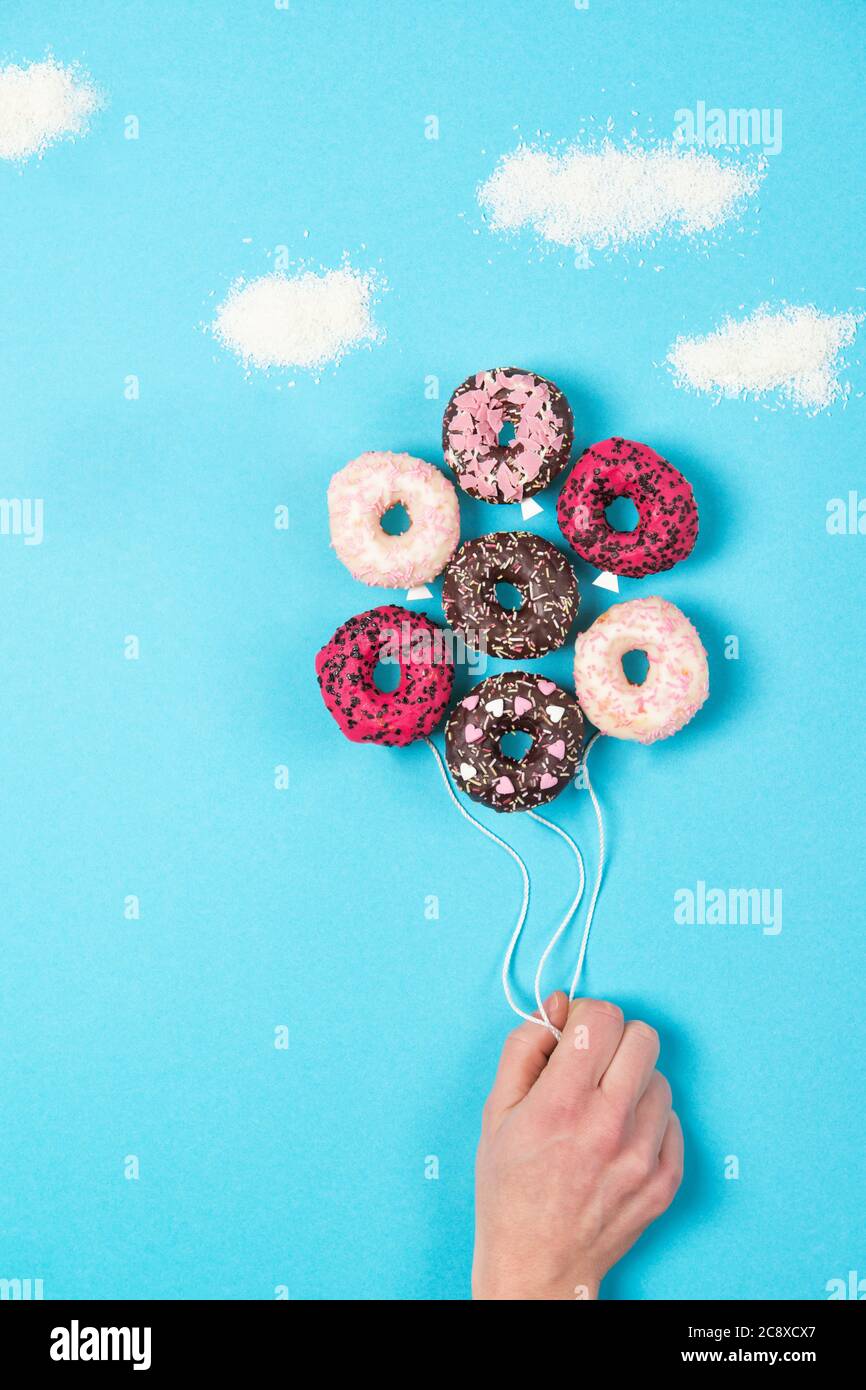 Mini beignets colorés sur fond bleu, idée de nourriture créative, main tenant des beignets en forme de ballons dans le ciel avec des nuages faits de noix de coco, vie supérieure Banque D'Images