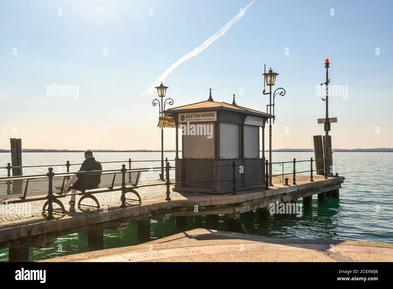 Vue à contre-jour d'un homme vu de derrière assis sur un banc de la jetée avec l'ancien kiosque de la billetterie du ferry, Lazise, Vérone, Vénétie, Italie Banque D'Images