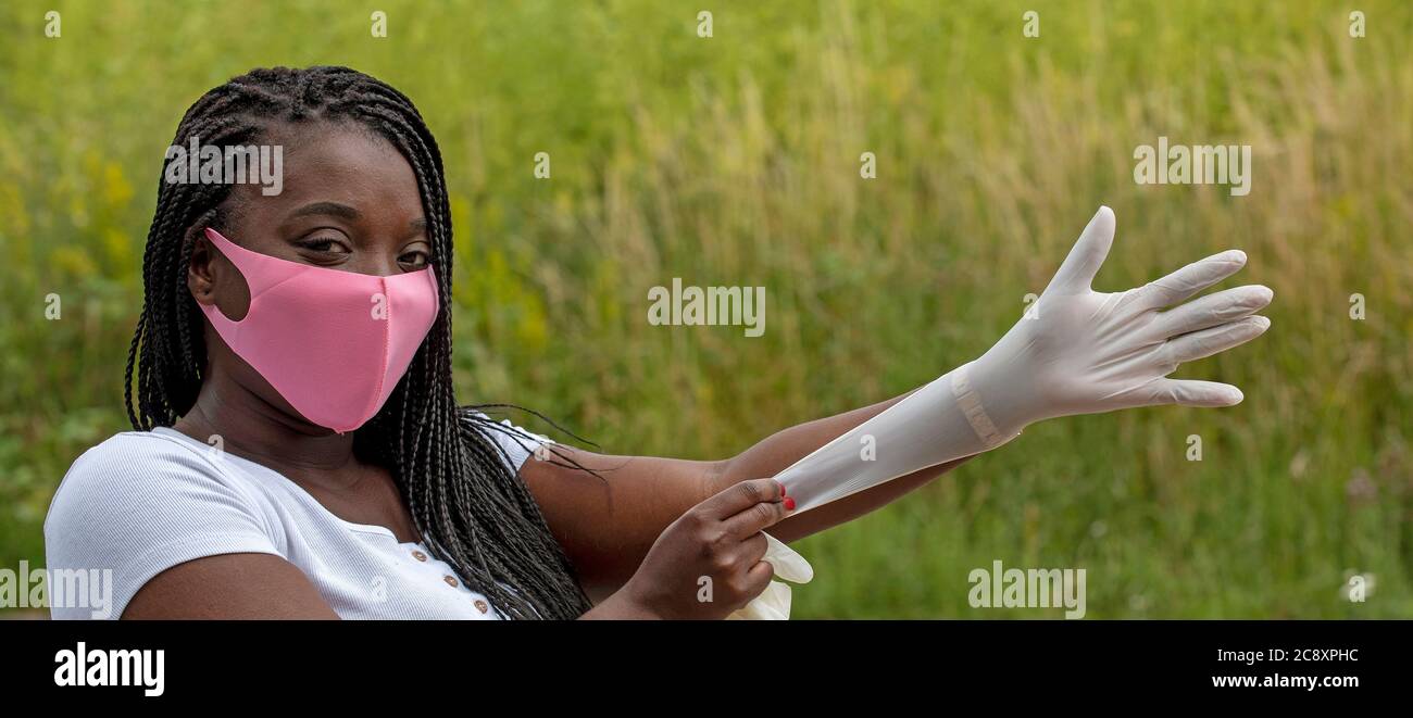 Angleterre, Royaume-Uni. 2020. Portrait d'une femme aux cheveux tressés portant un masque rose mettant des gants en caoutchouc pendant l'épidémie de Covid-19 Banque D'Images