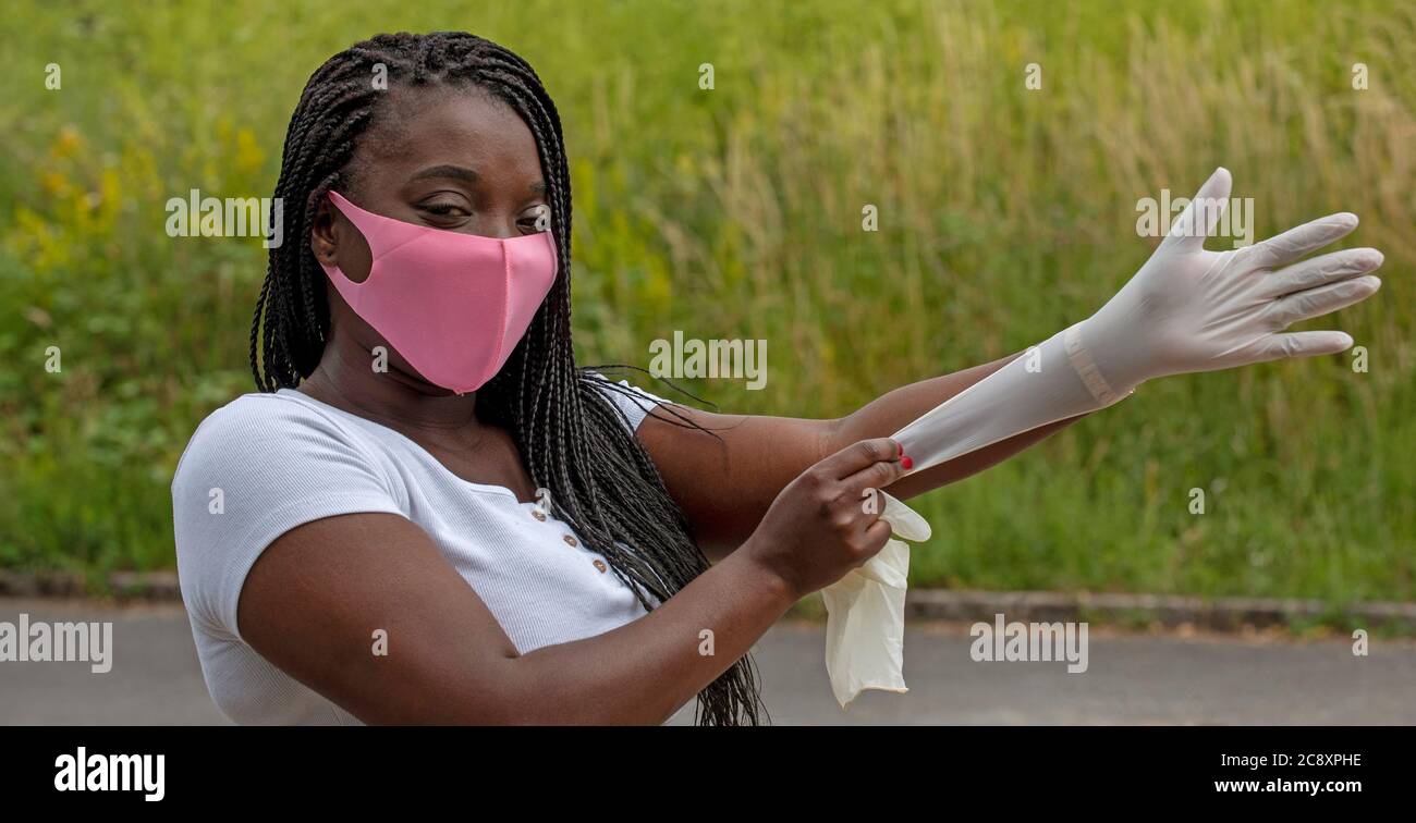 Angleterre, Royaume-Uni. 2020. Portrait d'une femme aux cheveux tressés portant un masque rose mettant des gants en caoutchouc pendant l'épidémie de Covid-19 Banque D'Images