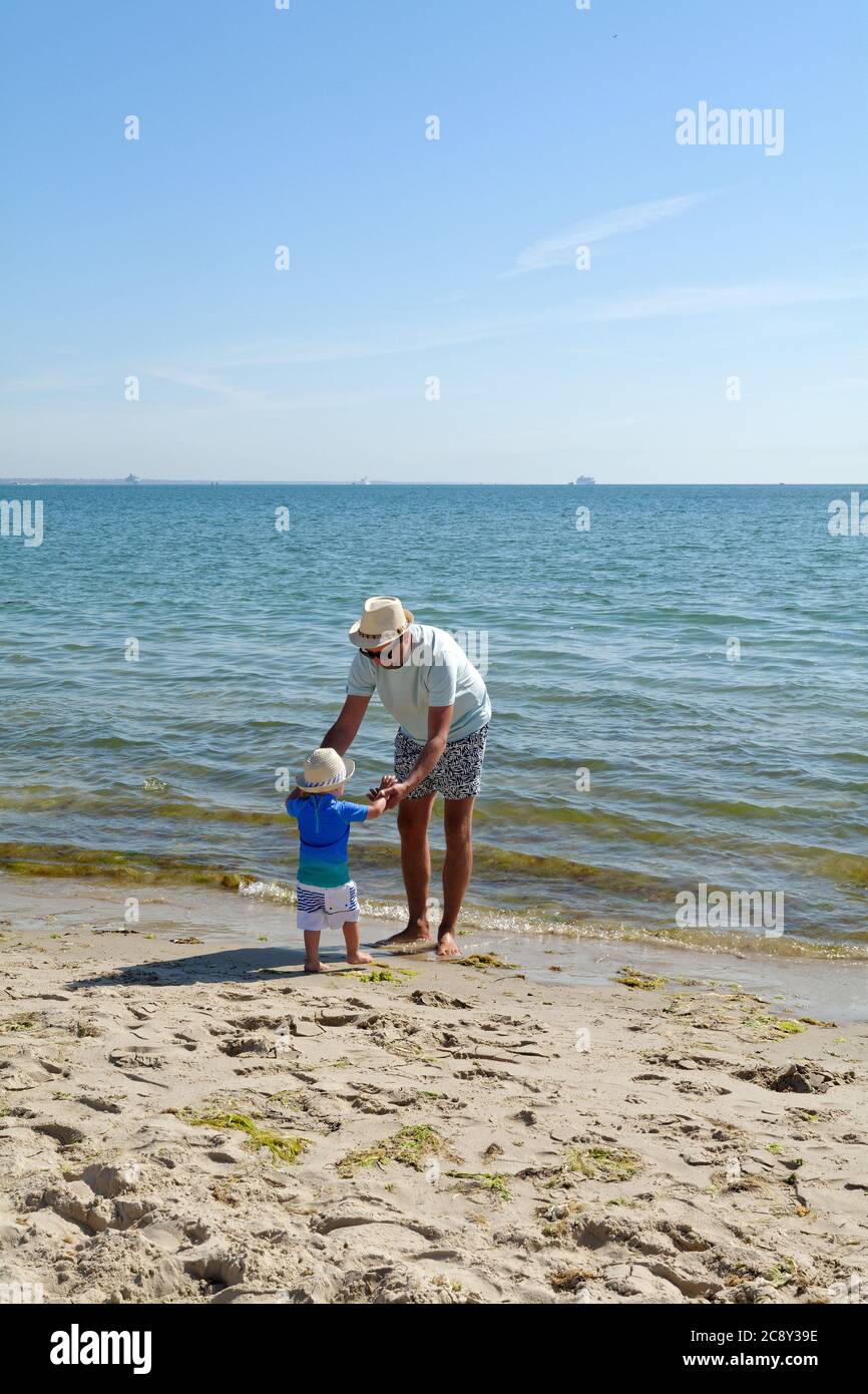 Un père et un fils tout-petits debout sur le bord de l'eau à la plage de Studland Bay, un jour d'été chaud, Dorset Angleterre Royaume-Uni Banque D'Images