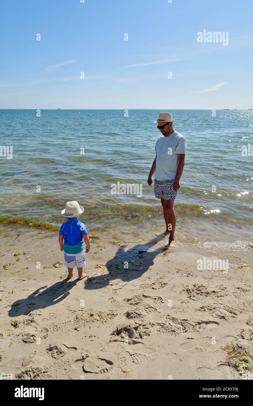 Un père et un fils tout-petits debout sur le bord de l'eau à la plage de Studland Bay, un jour d'été chaud, Dorset Angleterre Royaume-Uni Banque D'Images