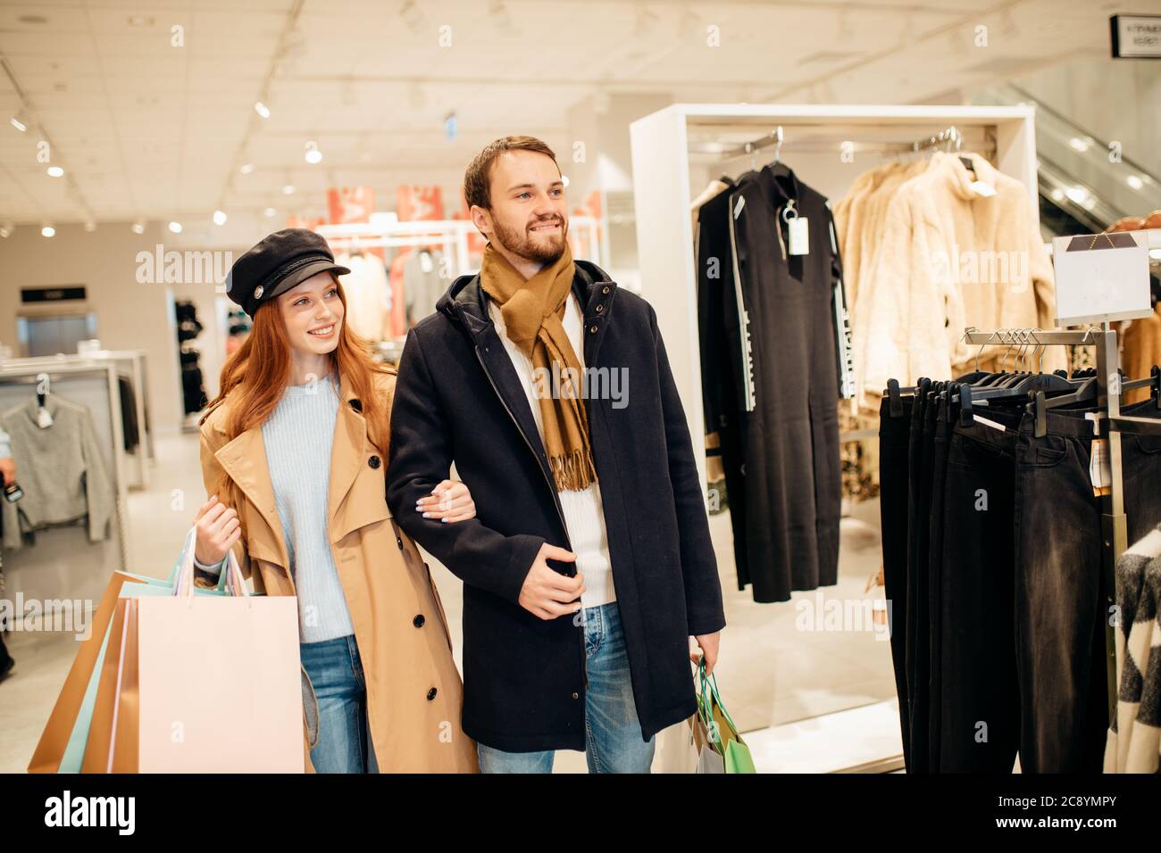 femme à poil rouge et homme à barbe caucasien en manteaux marcher dans le hall de la boutique de vêtements ensemble, choisir une nouvelle tenue Banque D'Images